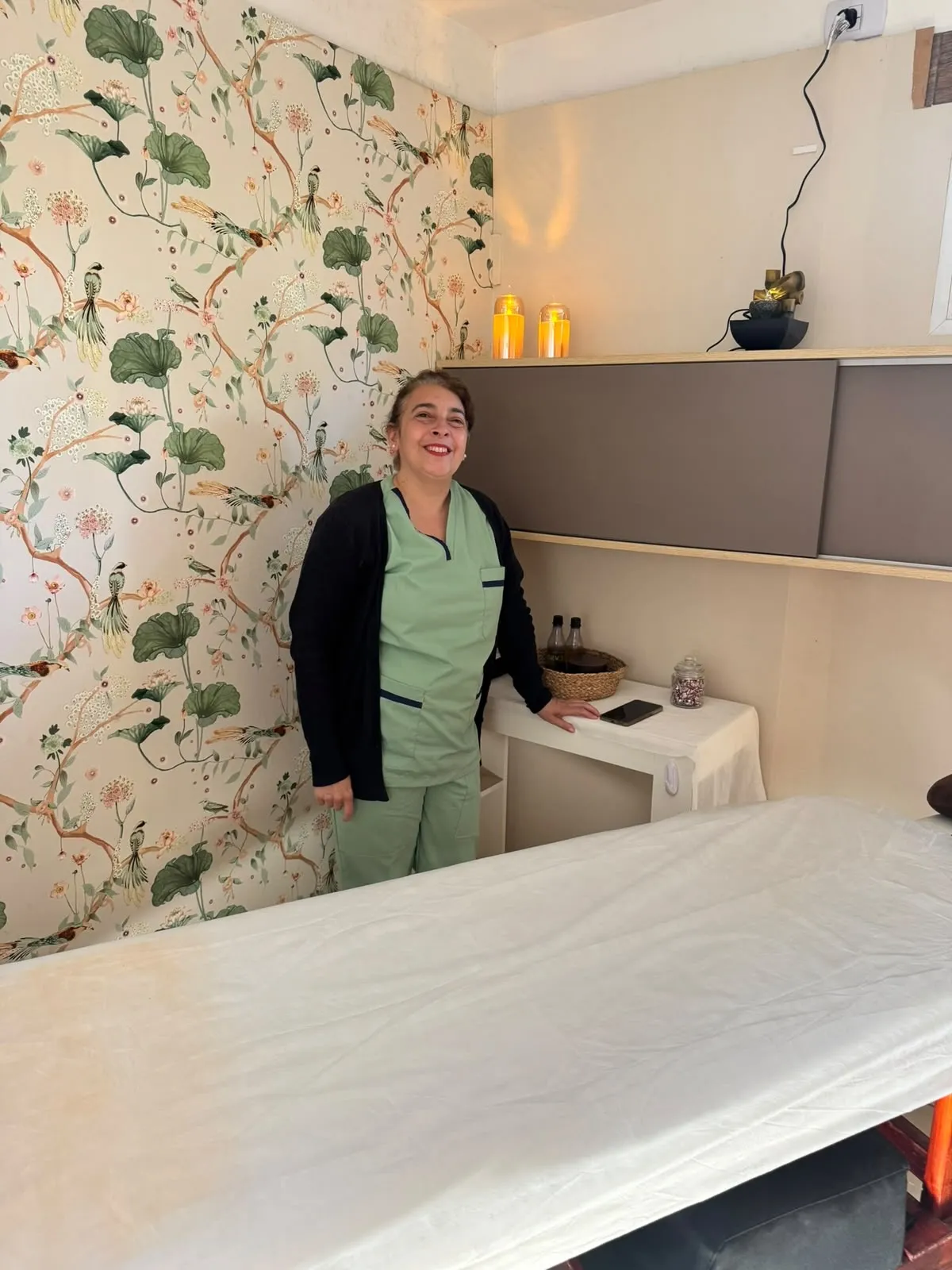 Sala de masajes de Benessere: Carolina, la masajista, junto a la camilla con sábana blanca, papel tapiz botánico y velas